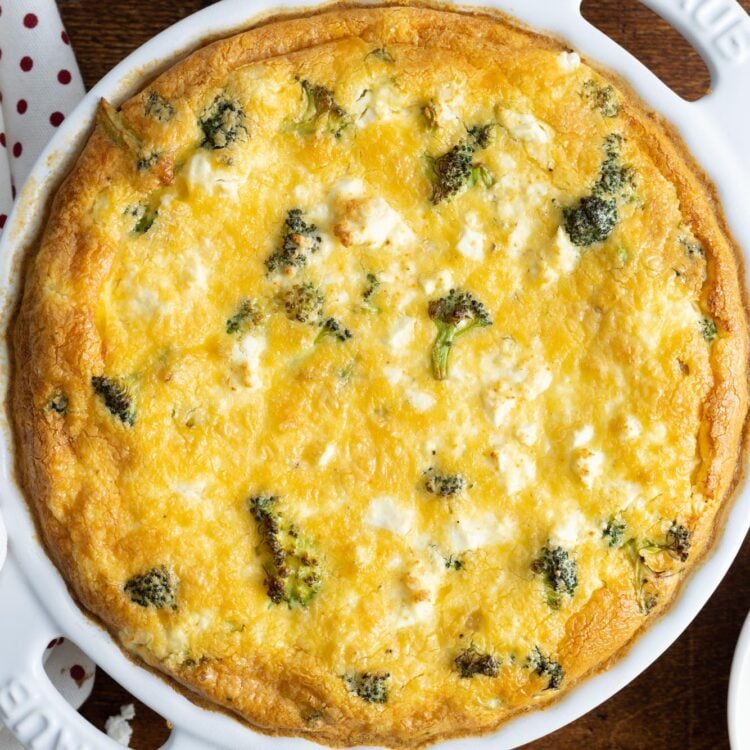 a crustless quiche with broccoli and feta cheese in a white ceramic pie dish. The quiche is set on a wooden table.