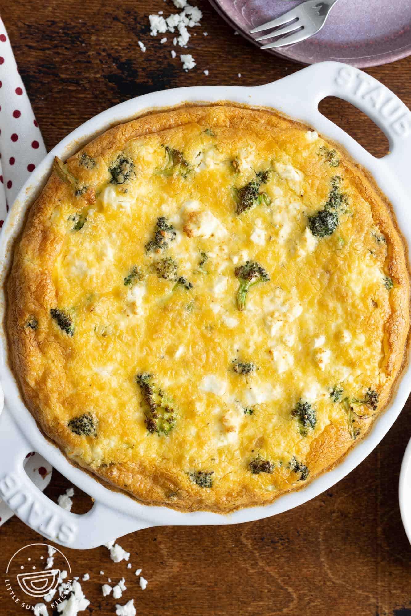 a crustless quiche with broccoli and feta cheese in a white ceramic pie dish. The quiche is set on a wooden table.