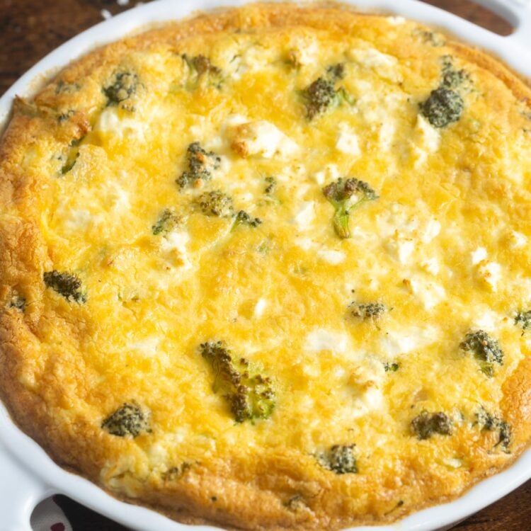 A whole crustless broccoli quiche on a wooden table. in the background are pink plates and small forks.