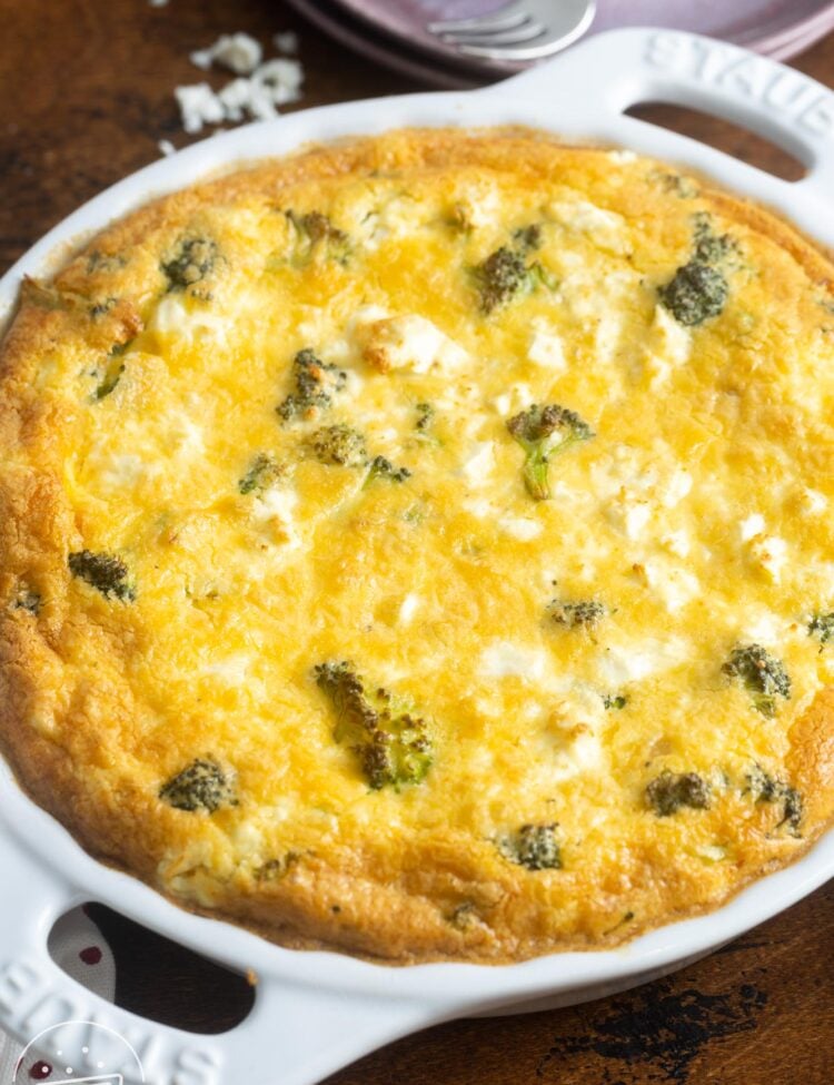 A whole crustless broccoli quiche on a wooden table. in the background are pink plates and small forks.