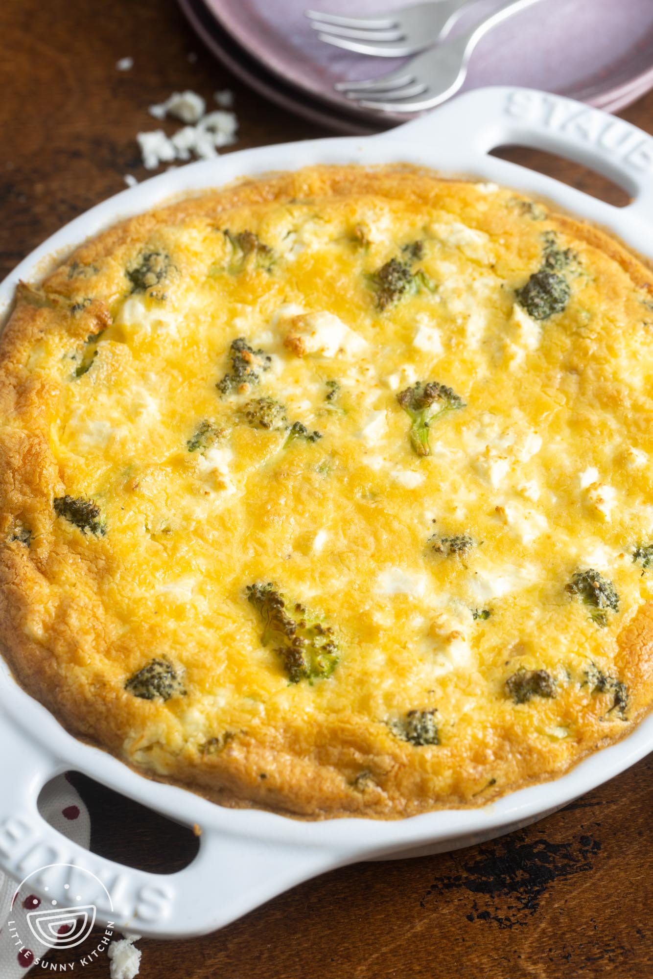 A whole crustless broccoli quiche on a wooden table. in the background are pink plates and small forks.