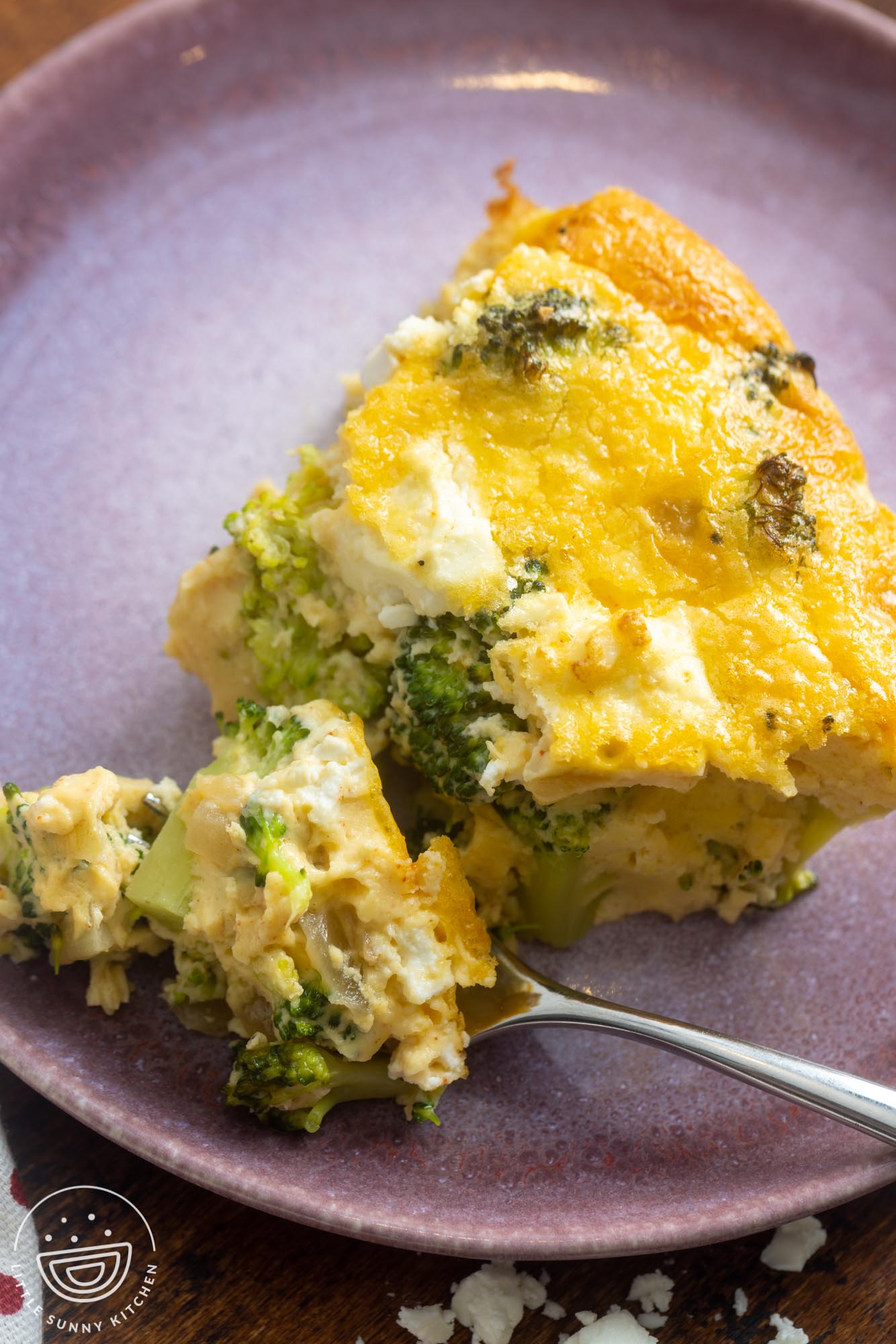 closeup of a slice of crustless broccoli quiche. A bite is on a fork, showing the creamy yet airy texture and the melty feta cheese.