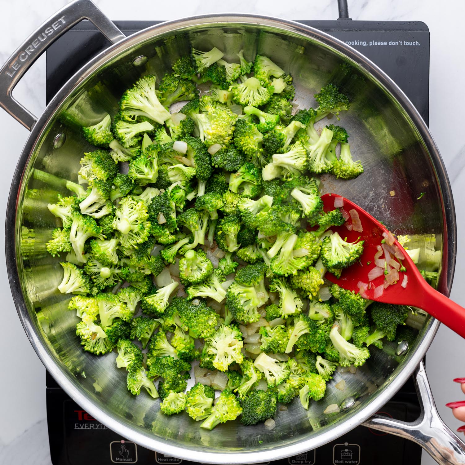 small broccoli florets being sauteed in a stainless steel skillet, stirred with a red spoon.