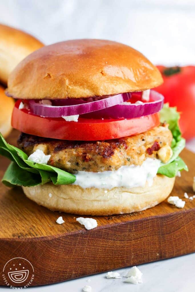 Grilled Greek chicken burger on a brioche bun with tzatziki, lettuce, tomato slices, red onion, and crumbled feta on a wooden board.