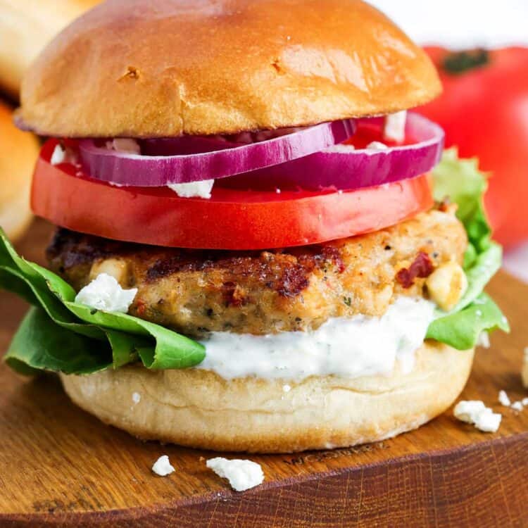 Grilled Greek chicken burger on a brioche bun with tzatziki, lettuce, tomato slices, red onion, and crumbled feta on a wooden board.