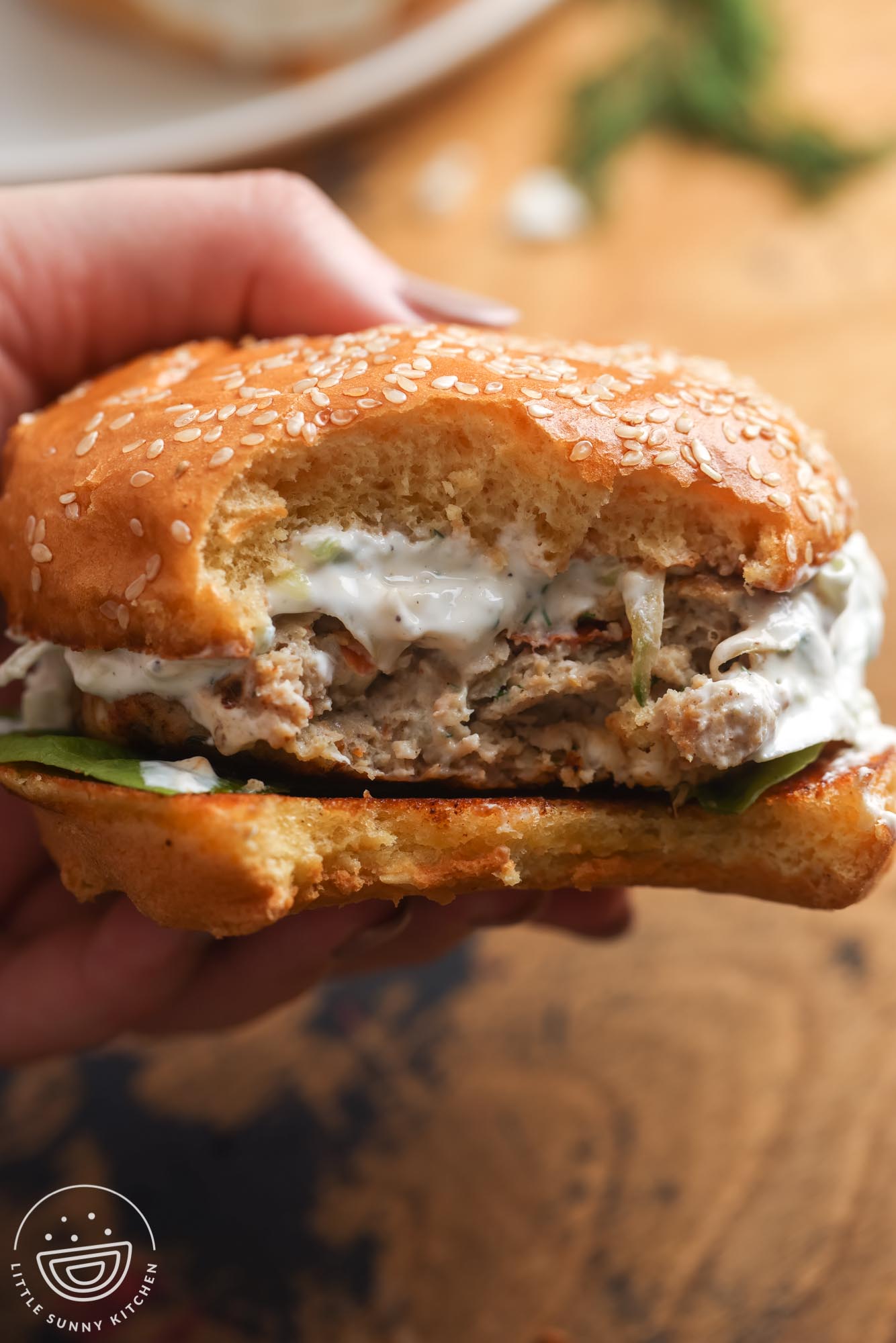 a hand holding a greek chicken burger with a creamy sauce on a bun.