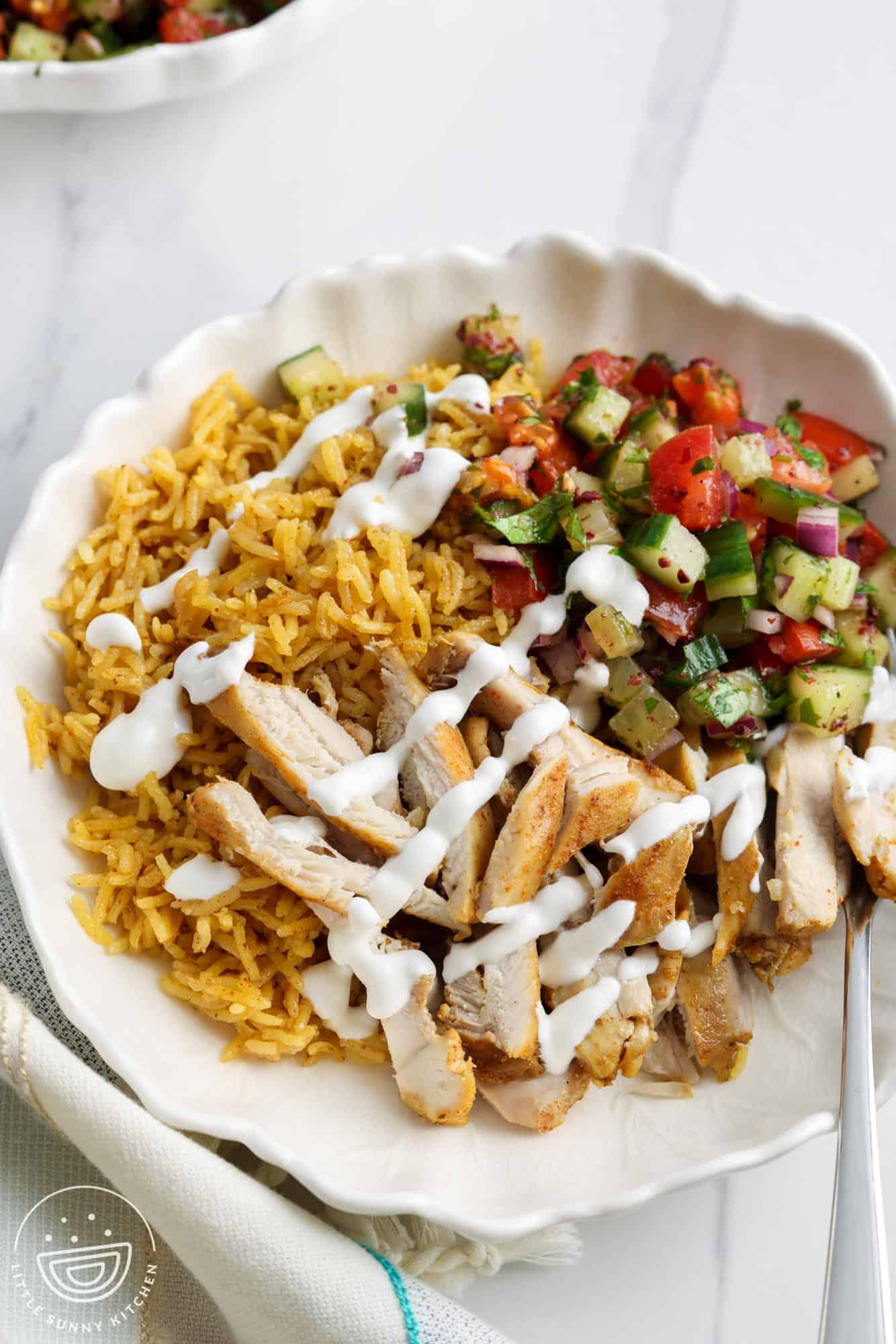 a bowl of shawarma rice with sliced chicken and cucumber shirazi salad, drizzled with yogurt sauce.