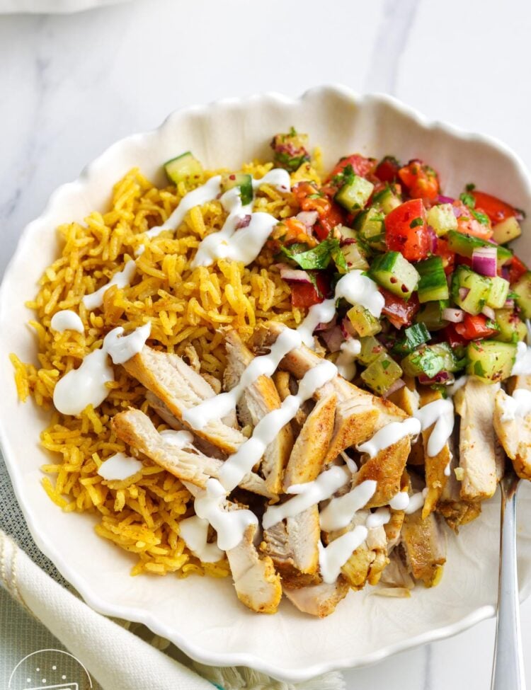 a bowl of shawarma rice with sliced chicken and cucumber shirazi salad, drizzled with yogurt sauce.