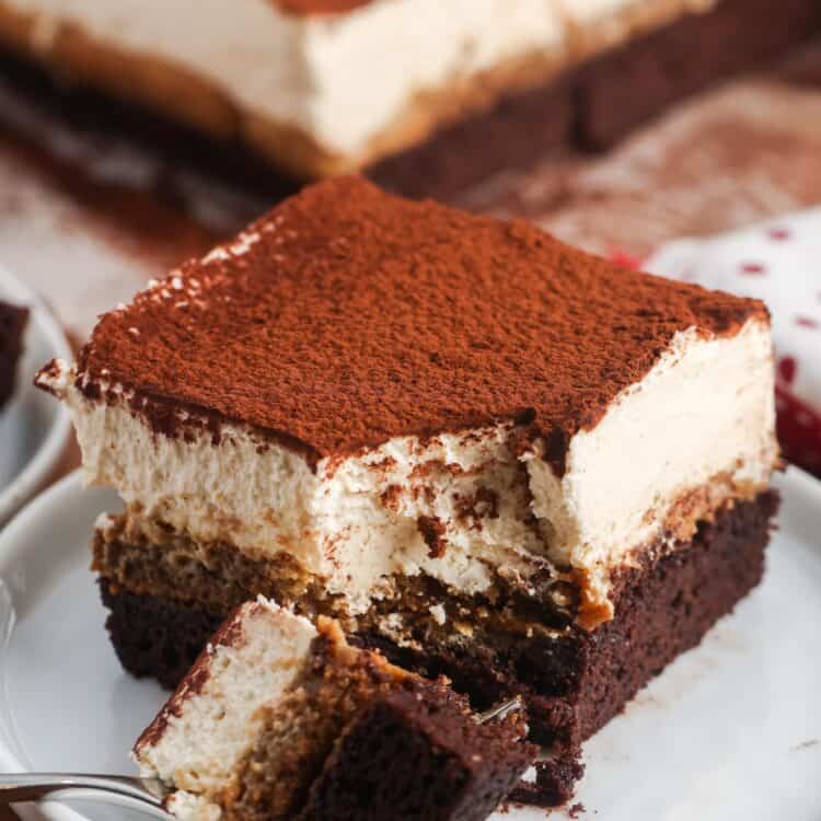 a tiramisu brownie on a small white dessert plate. A fork has removed a bite from the front corner, showing the creamy texture of the topping.