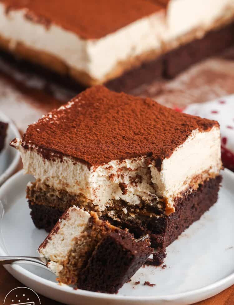 a tiramisu brownie on a small white dessert plate. A fork has removed a bite from the front corner, showing the creamy texture of the topping.