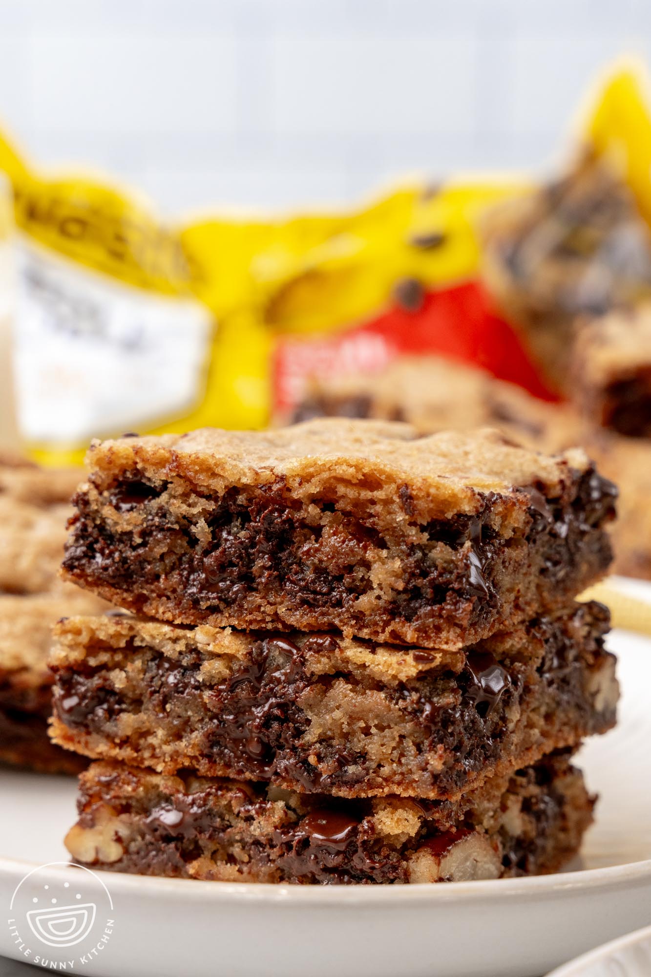 three toll house cookie bars, stacked, on a white plate. In the background is a bag of chocolate chips.