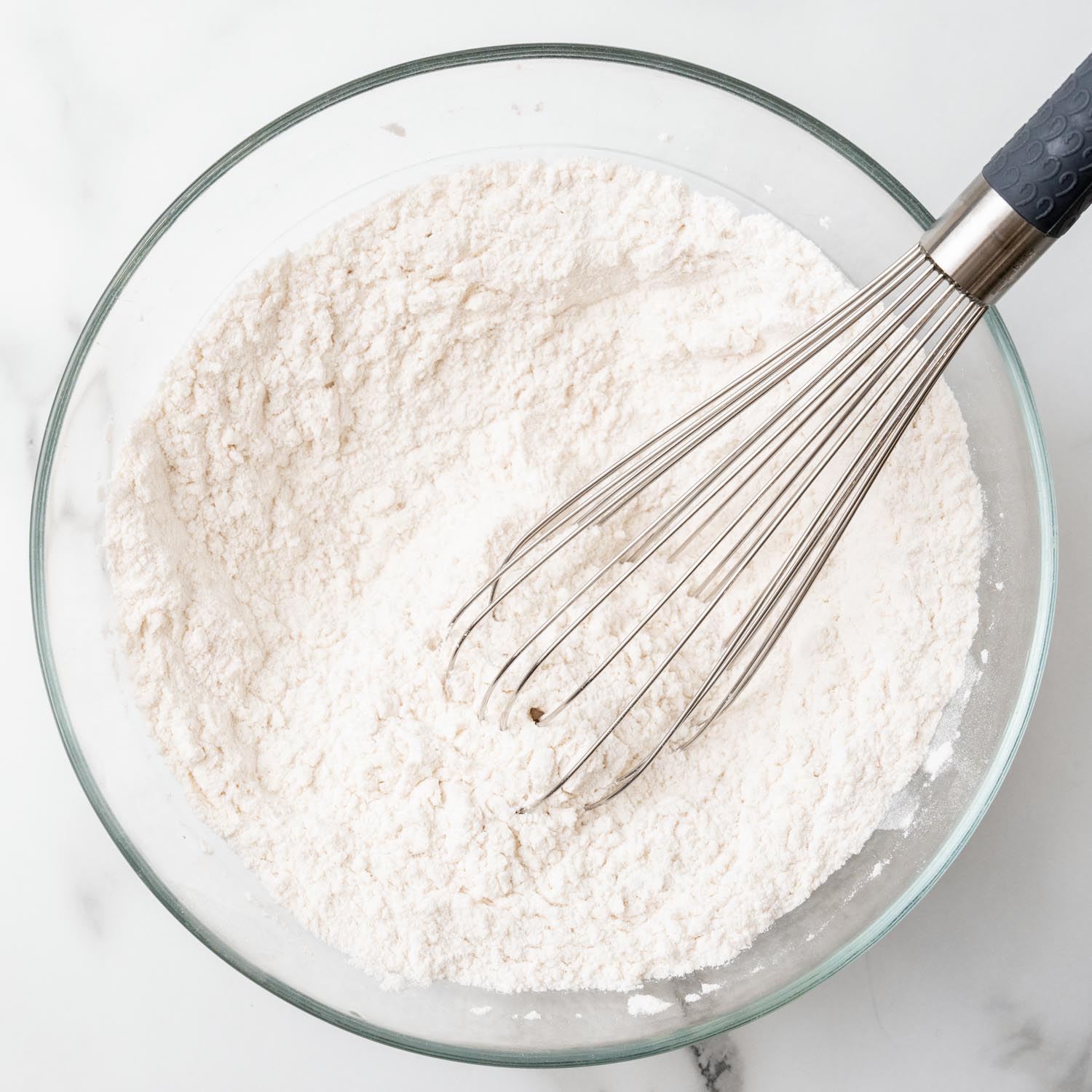 A glass bowl of flour, stirred with a whisk.