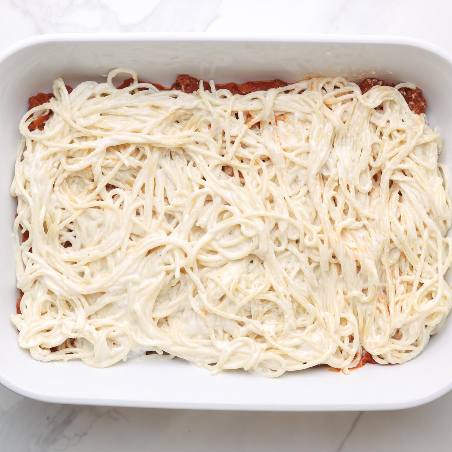 cream cheese spaghetti added to a casserole dish on top of tomato meat sauce.