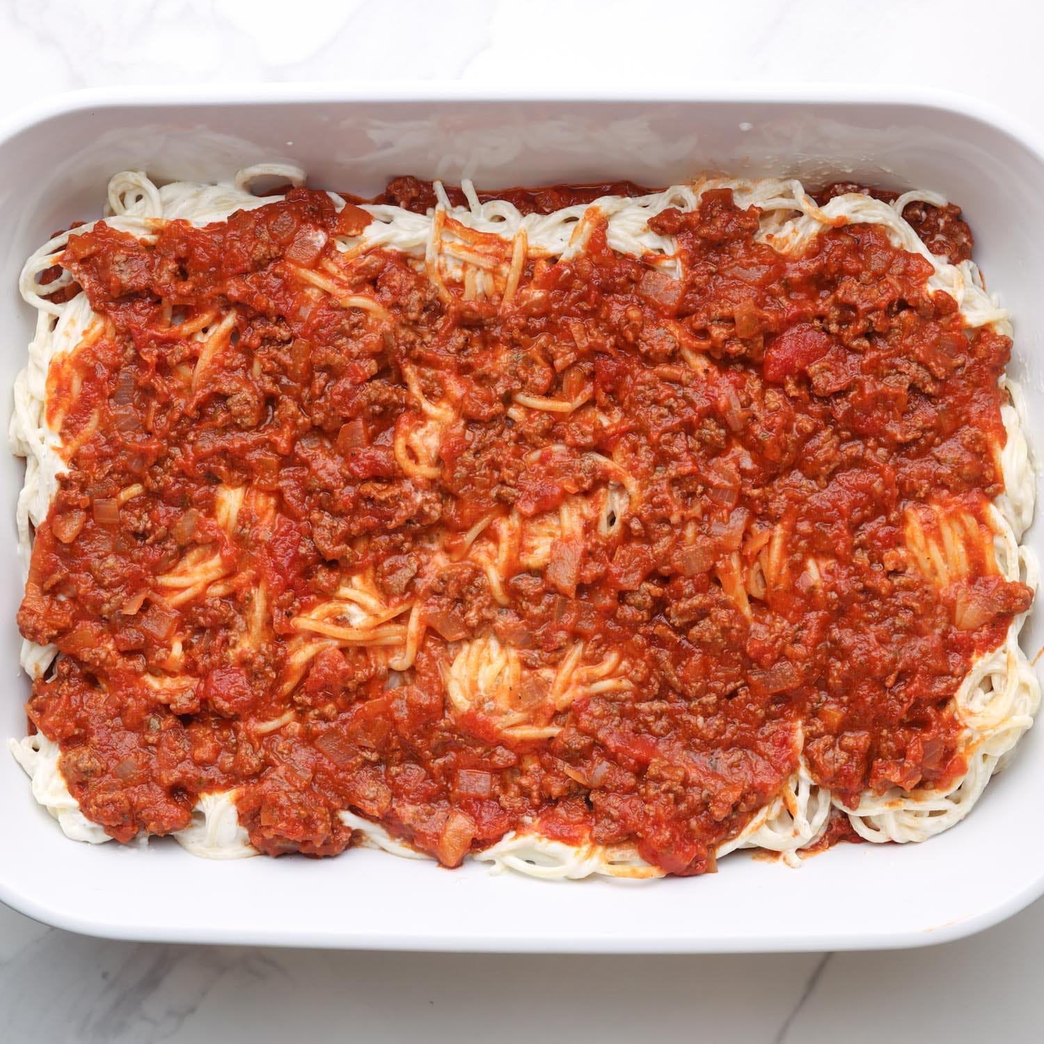 meat sauce added on top of cream cheese spaghetti in a white casserole dish.