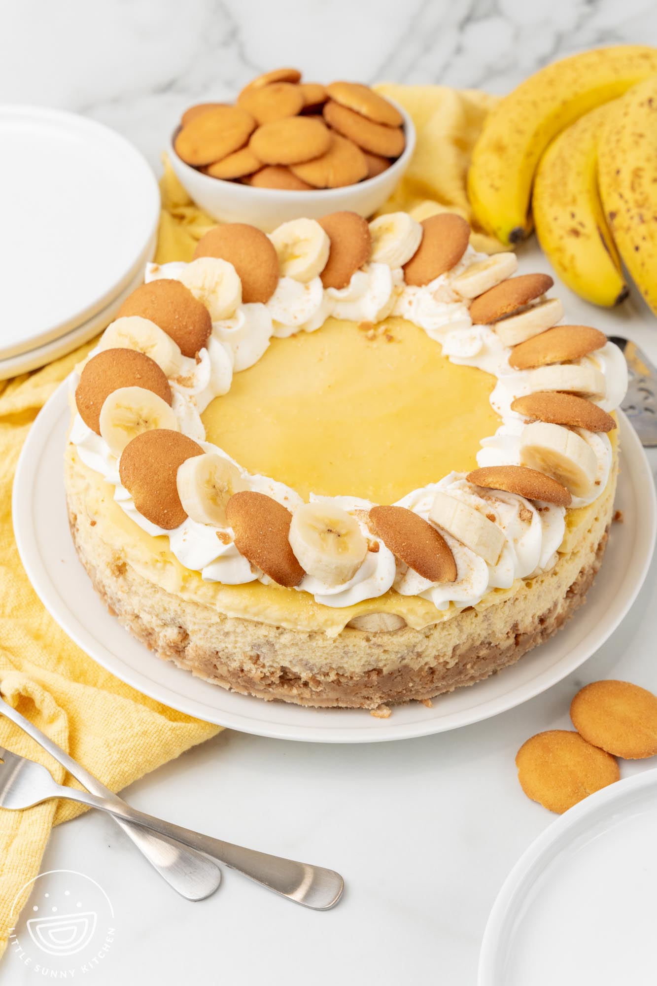 A whole banana pudding cheesecake with vanilla wafer crust and sliced bananas on top, served on a round white platter. behind the cheesecake is a bowl of vanilla wafers and a bunch of ripe bananas.