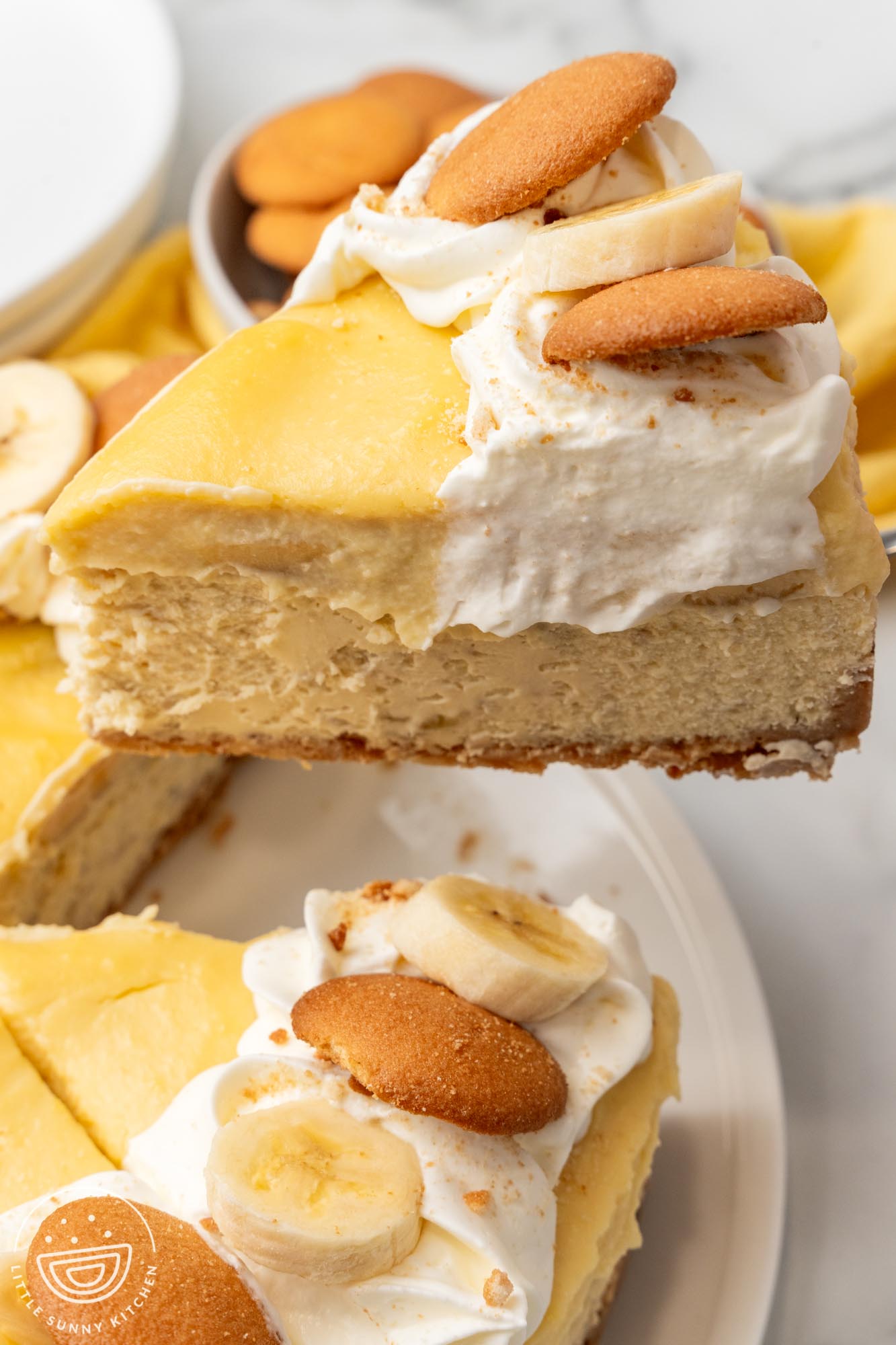 A large slice of banana pudding cheesecake lifted up from a whole cheesecake. The pieces are topped with whipped cream and vanilla wafers.