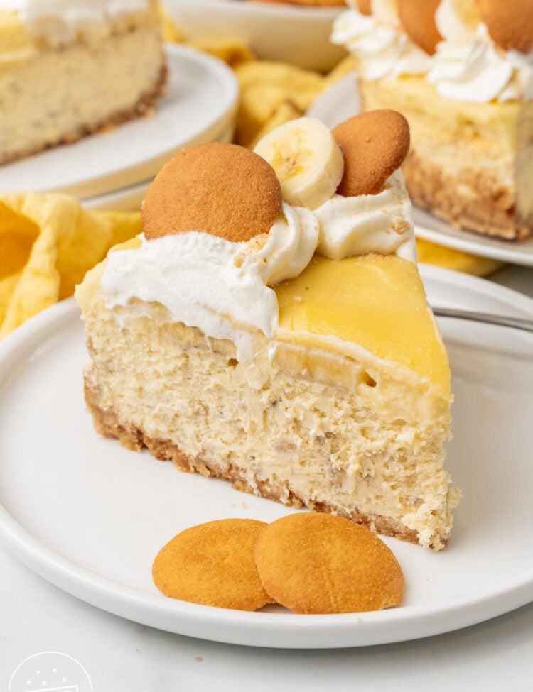 A slice of banana pudding cheesecake on a white dessert plate with two vanilla wafers. The pie is topped with whipped cream, cookies, and fresh banana slices.