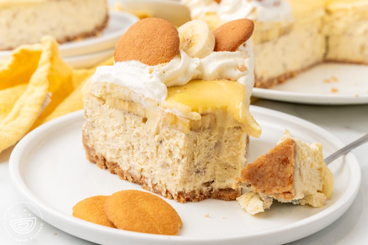 A slice of banana pudding cheesecake on a plate with two vanilla wafer cookies. A fork has taken a bite from the tip of the slice.