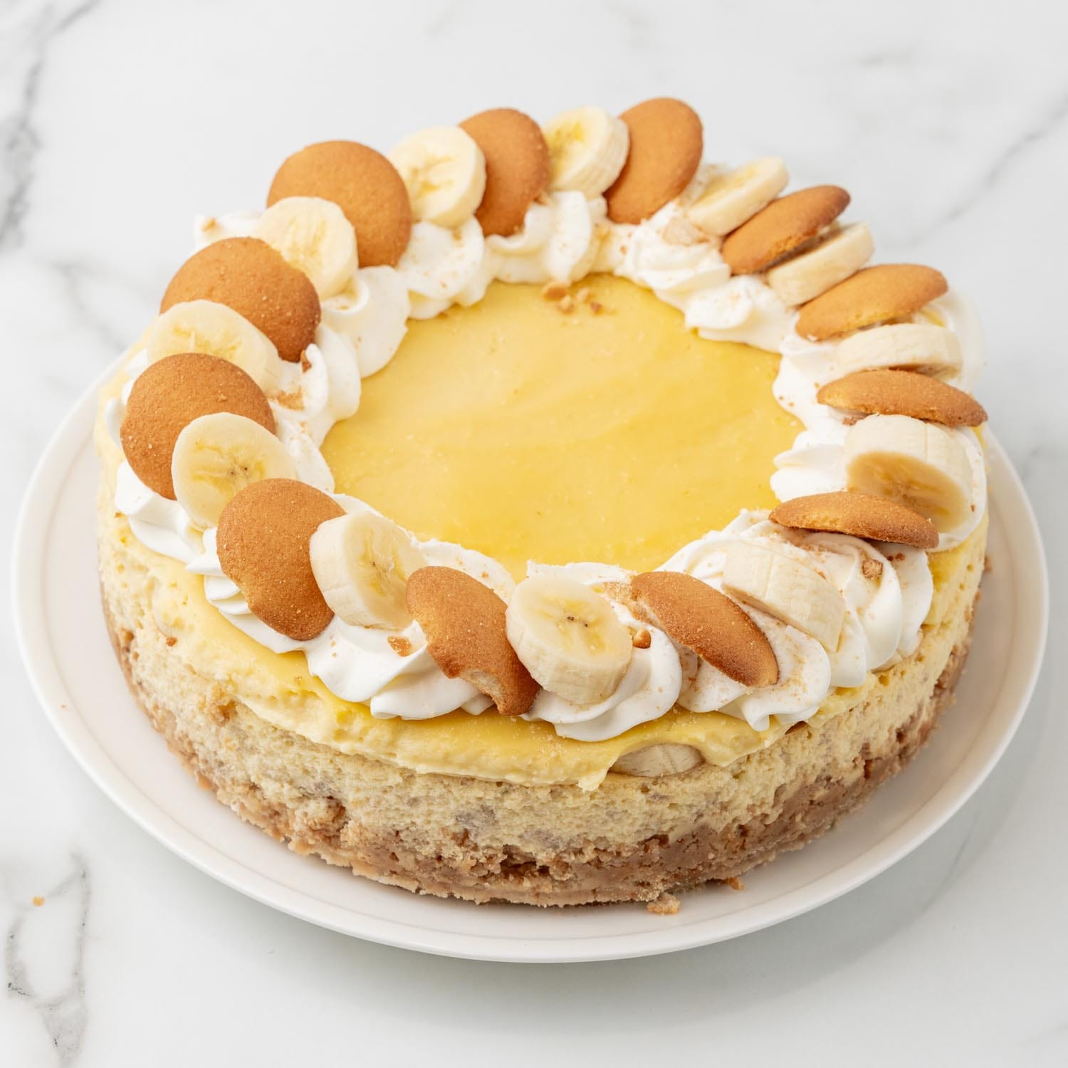 A round cheesecake topped with a ring of whipped cream, sliced bananas, and vanilla wafer cookies.