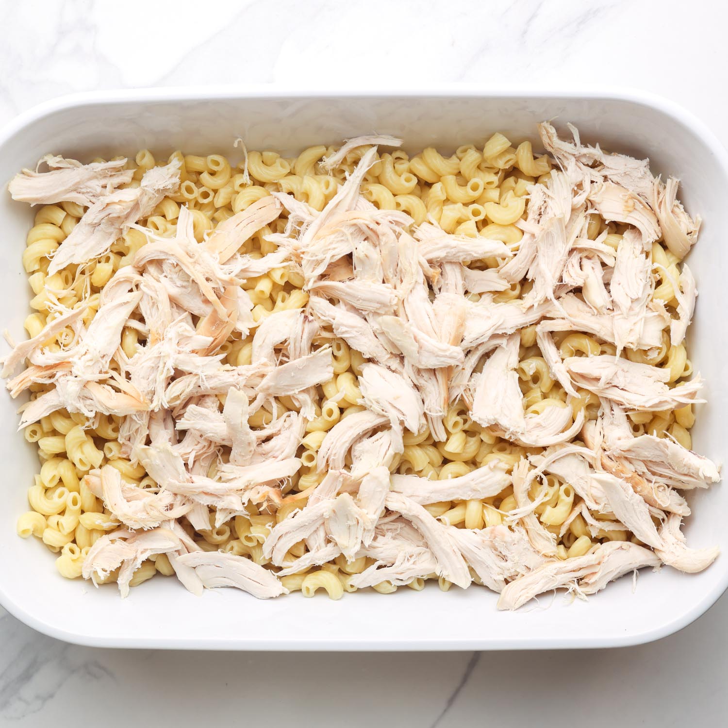 shredded chicken added to cooked macaroni in a rectangular baking pan.