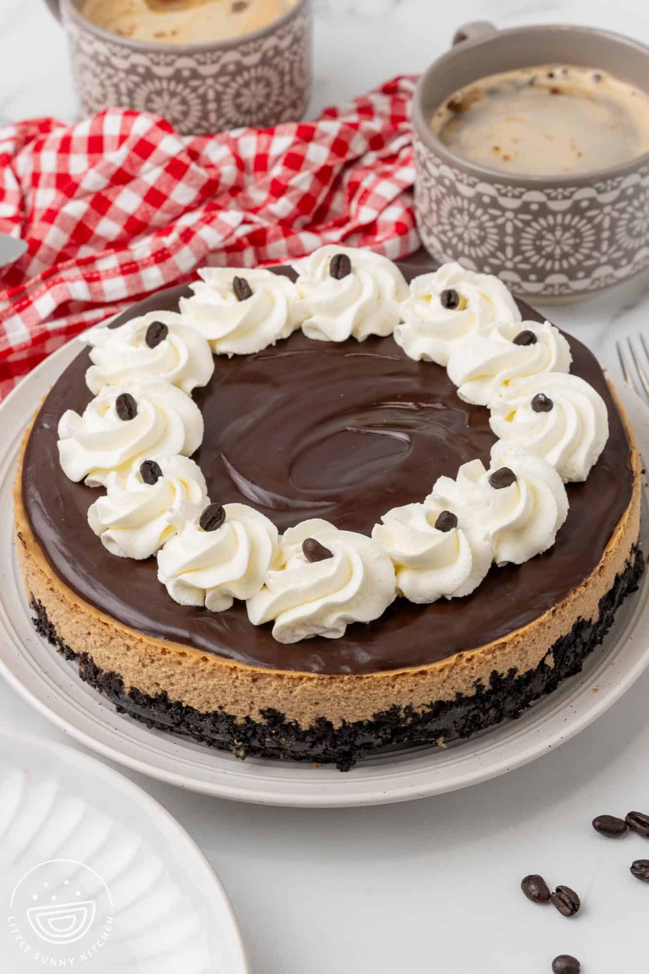 homemade coffee cheesecake with oreo crust, topped with ganache, whipped cream, and coffee beans on a large plate. Behind the plate are two gray patterned mugs of coffee and a white and red checkered kitchen towel.