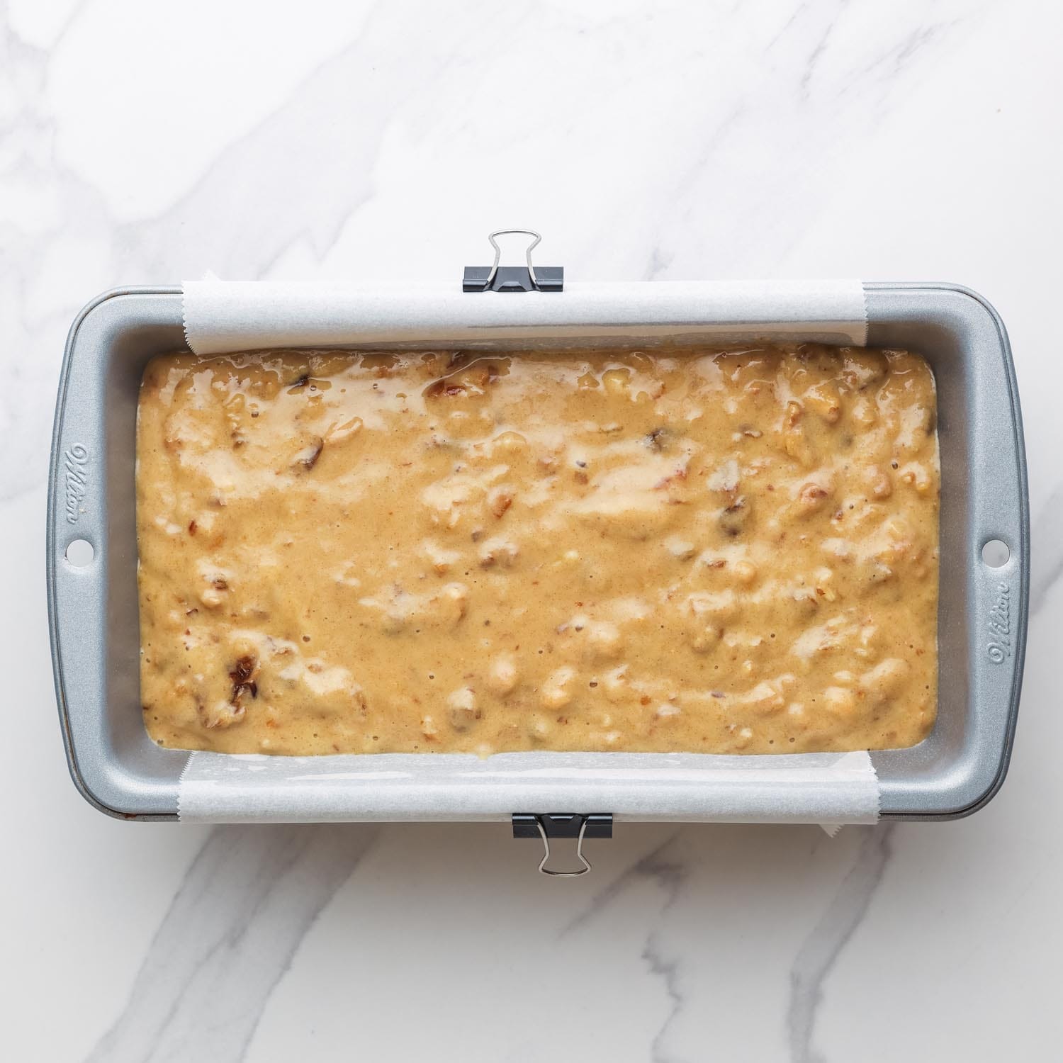 date nut bread batter added to a loaf pan lined with parchment paper. The paper is held in place with metal binder clips.