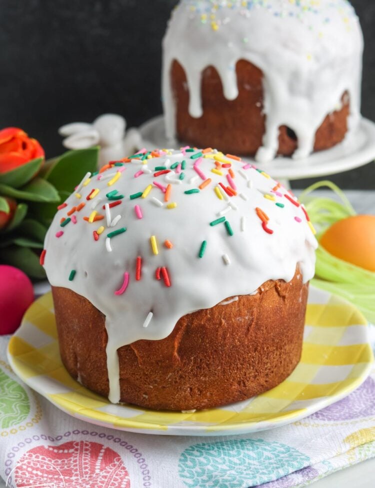 Kulich loaves on small plates on top of an easter egg decorated towel with flowers in the background.