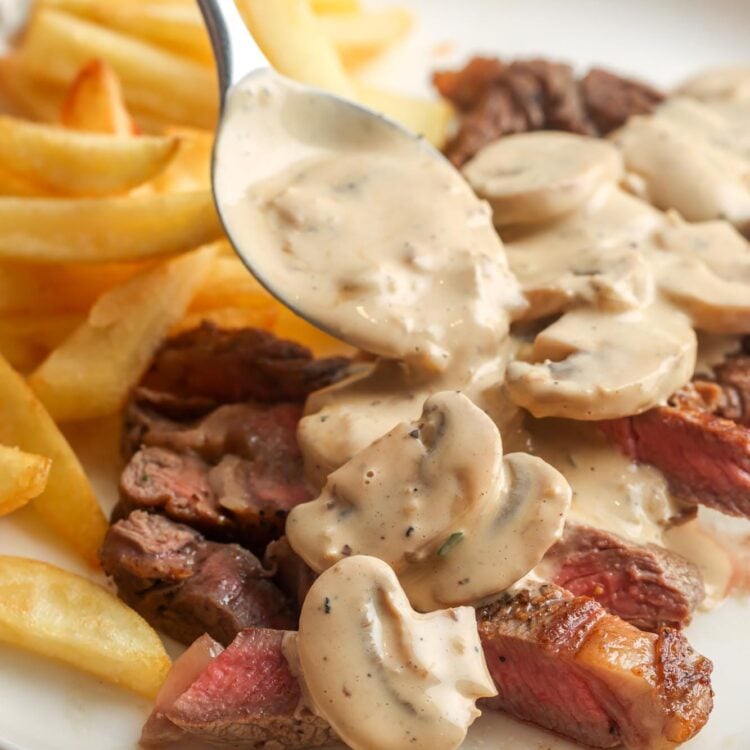 A plate of sliced steak and french fries with a creamy sauce with mushrooms spooned over.