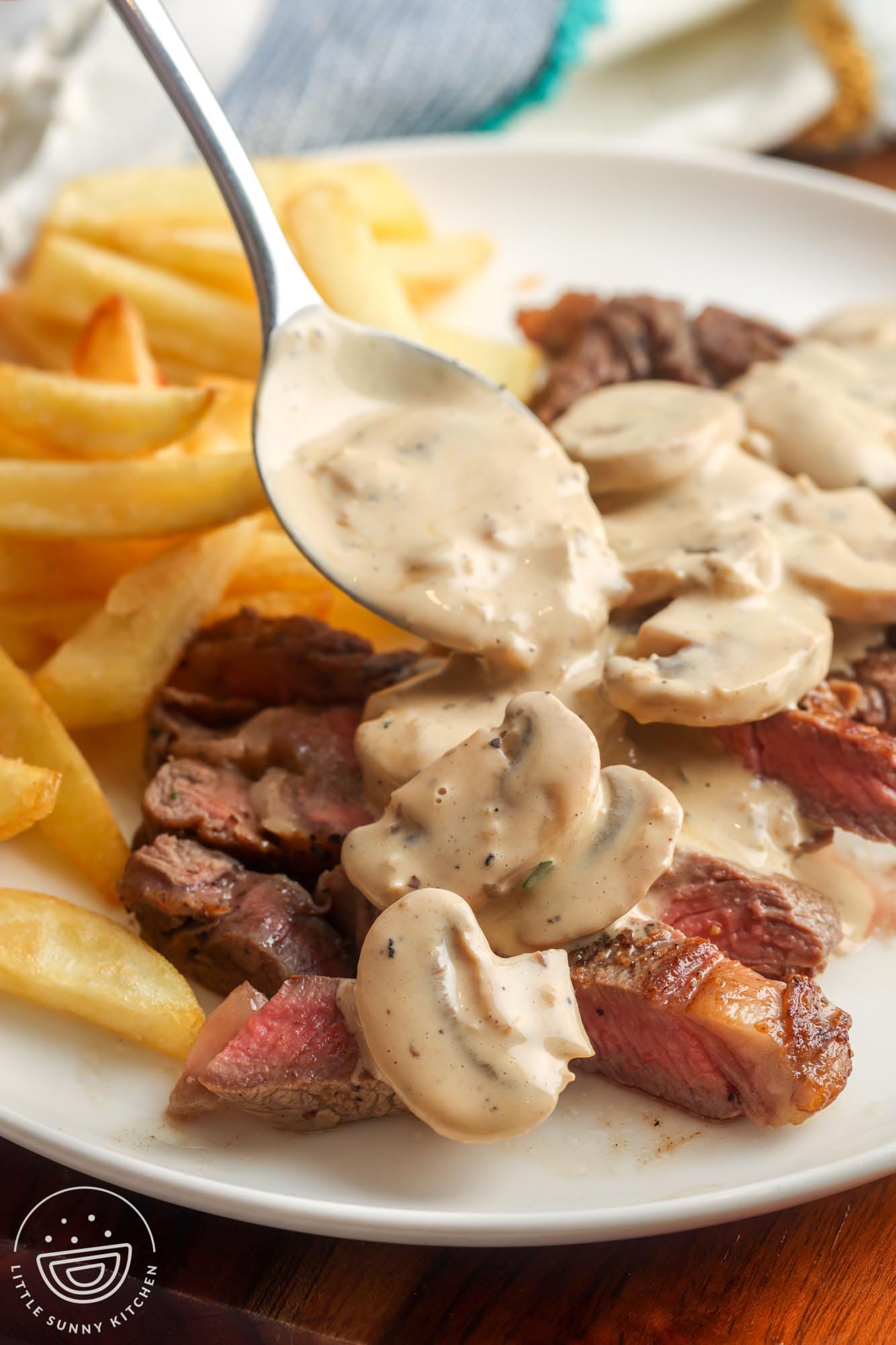 A plate of sliced steak and french fries with a creamy sauce with mushrooms spooned over.