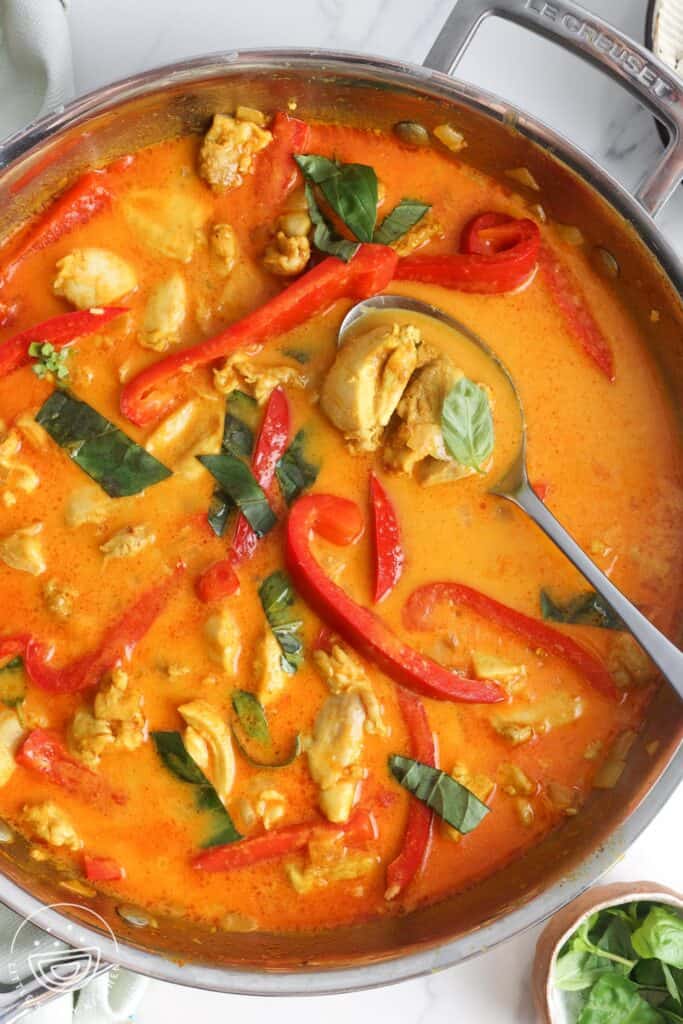 A stainless steel skillet of Thai red curry with chicken and red bell peppers, garnished with Thai basil leaves.