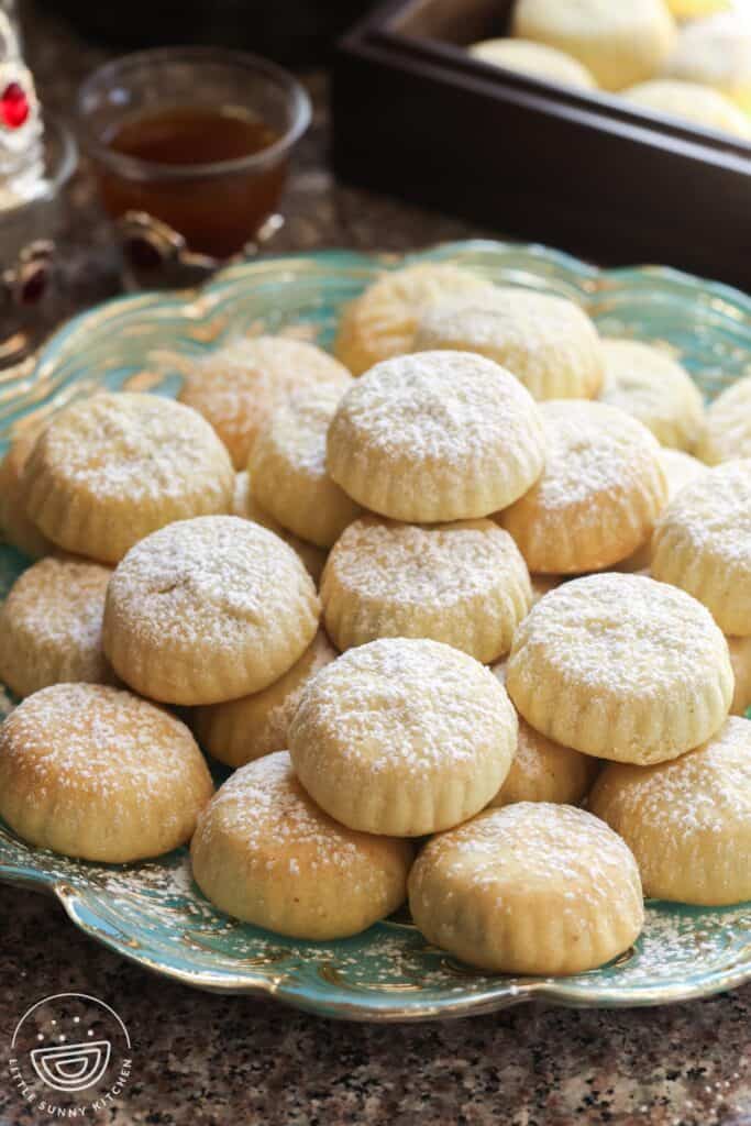 Stacked maamoul cookies with a soft semolina crust, one showing a bite revealing the dark date filling, lightly dusted with powdered sugar.