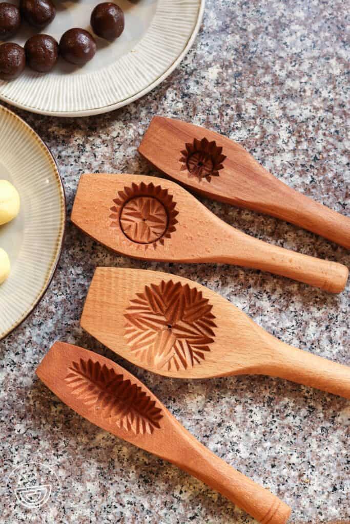 Wooden maamoul molds with intricate carved designs laid on a countertop, beside plates of date paste balls and semolina dough ready to be shaped.