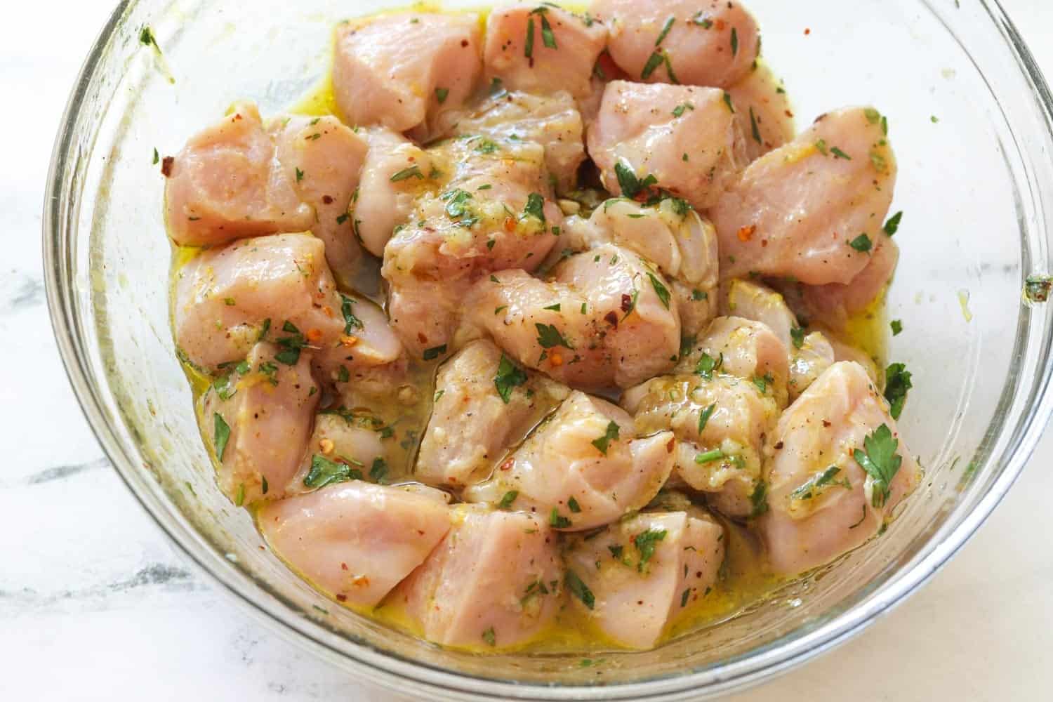 Chicken cubes marinating in oil, garlic, and seasonings in a glass bowl.