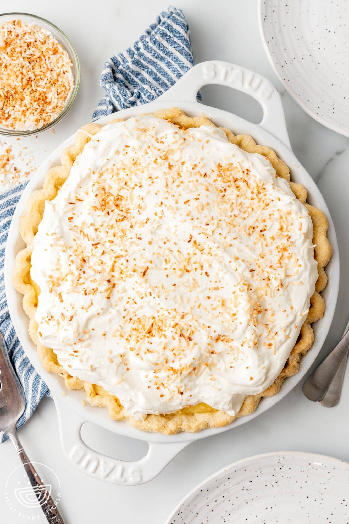 overhead view of a whole coconut cream pie. Next to the pie are two plates, a blue striped towel, and a small bowl of browned toasted shredded coconut.
