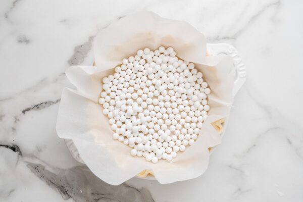 parchment paper and pie weights added to pie crust in a white ceramic dish. The pie crust is set on a white marble surface.