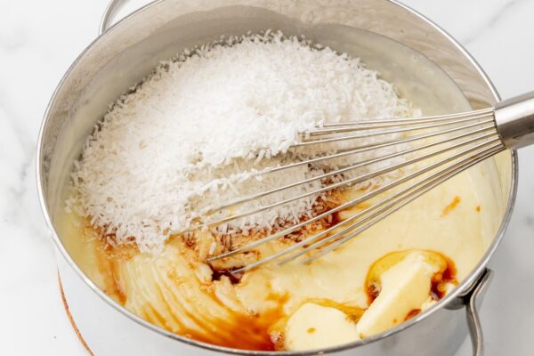 butter, vanilla, and sweetened coconut whisked into the filling for coconut cream pie in a metal saucepan.