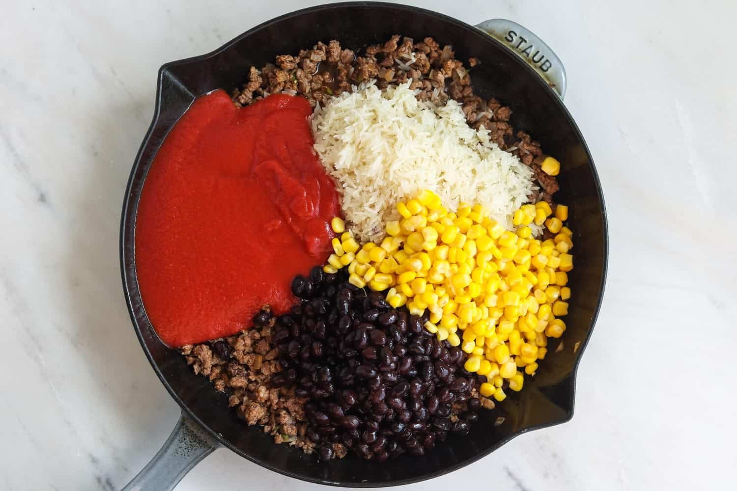 a cast iron skillet with cooked ground beef, uncooked rice, corn, black beans, and tomato sauce.