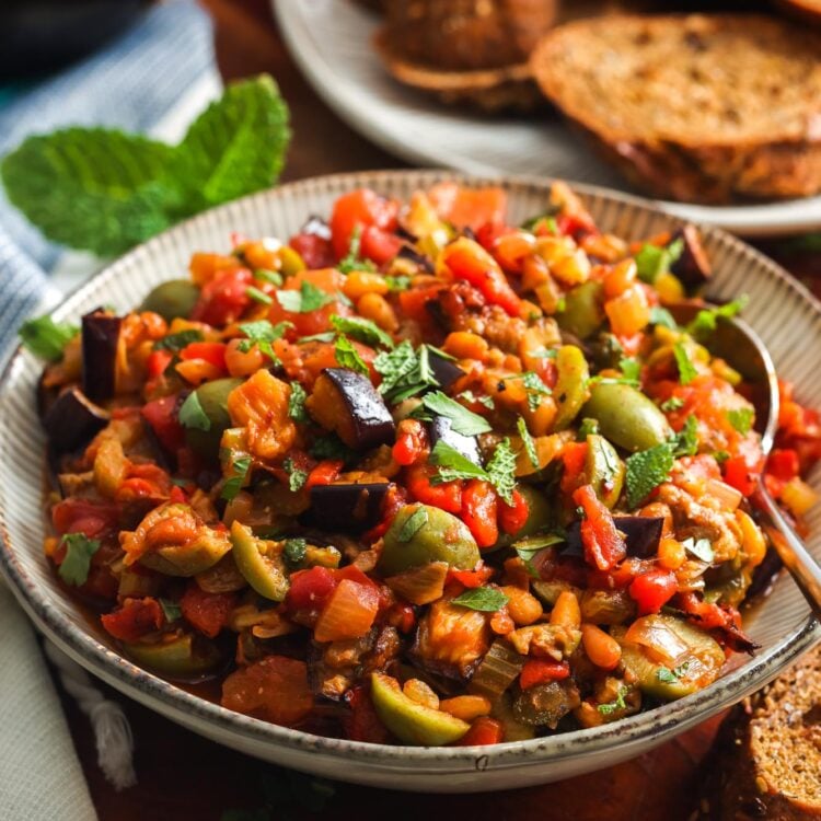 a bowl of homemade caponata on a wooden table next to a platter of whole grain crostini.