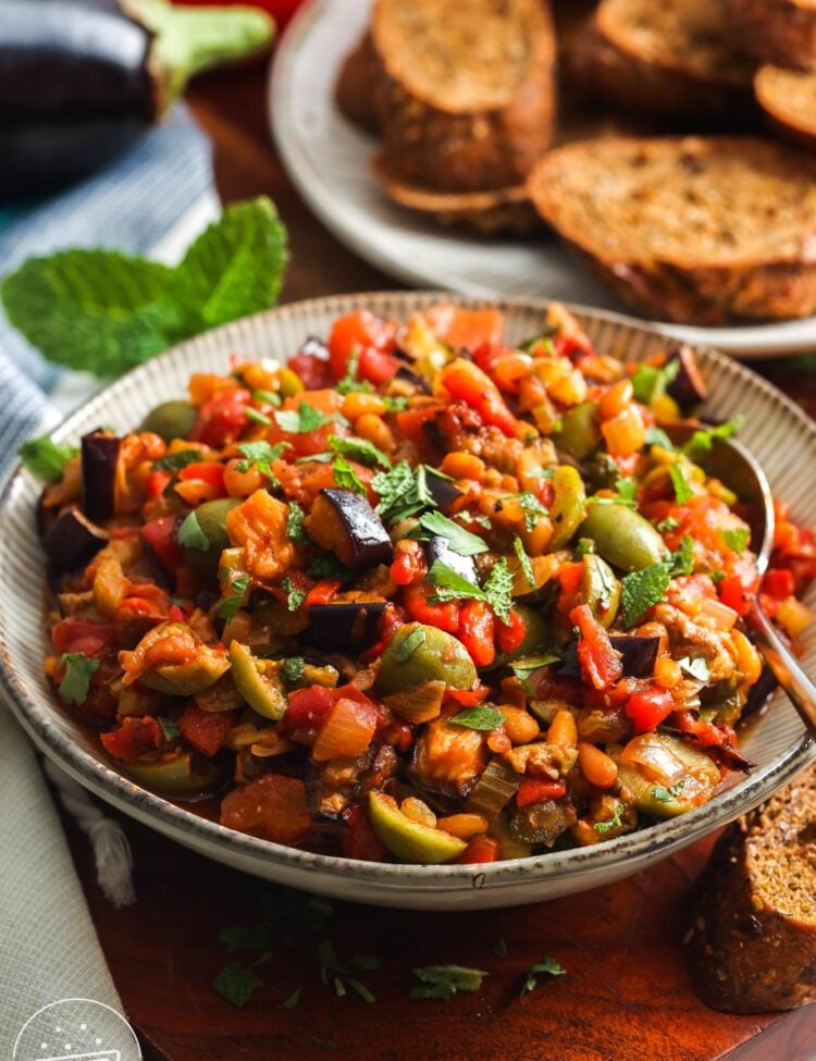 a bowl of homemade caponata on a wooden table next to a platter of whole grain crostini.