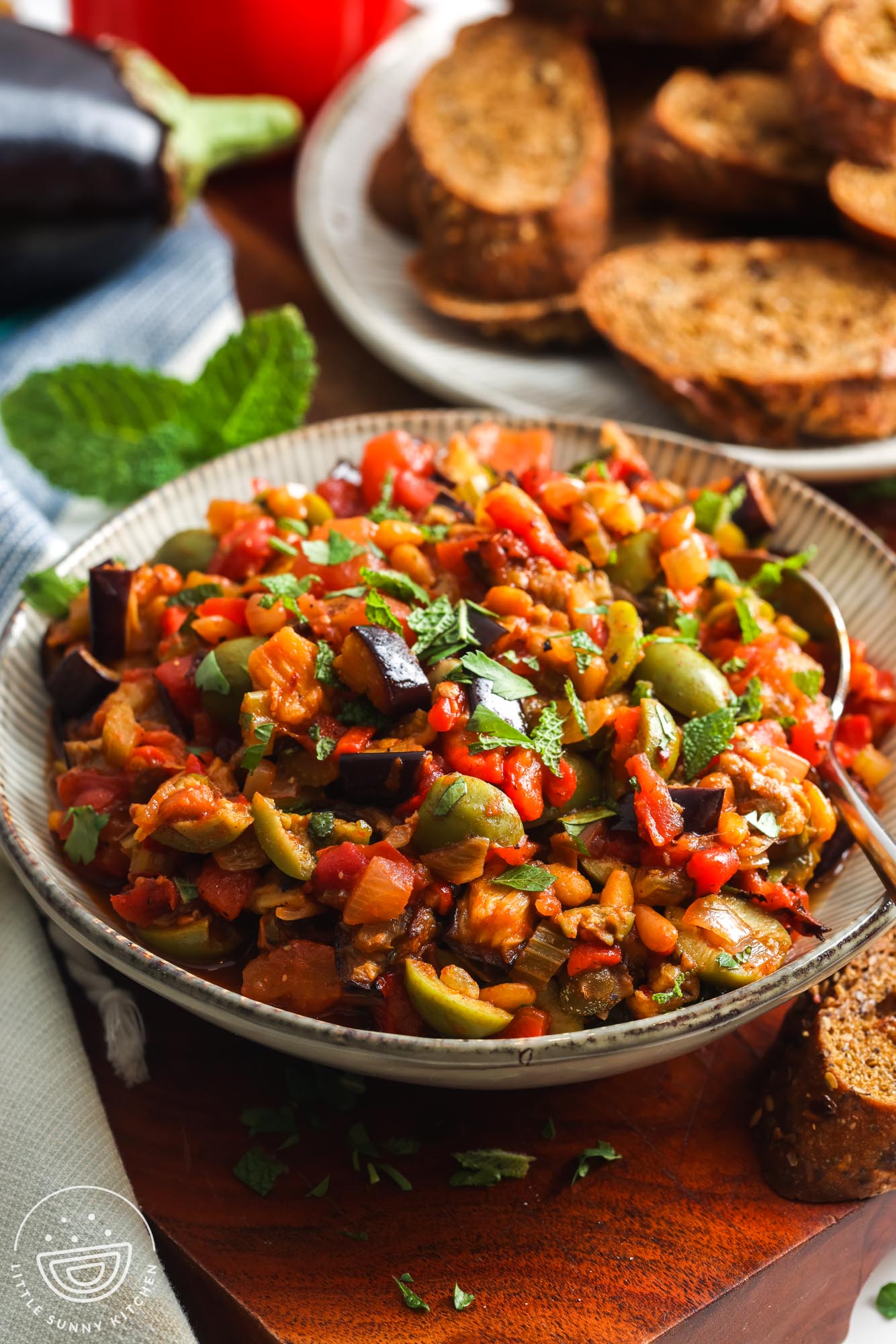 a bowl of homemade caponata on a wooden table next to a platter of whole grain crostini.