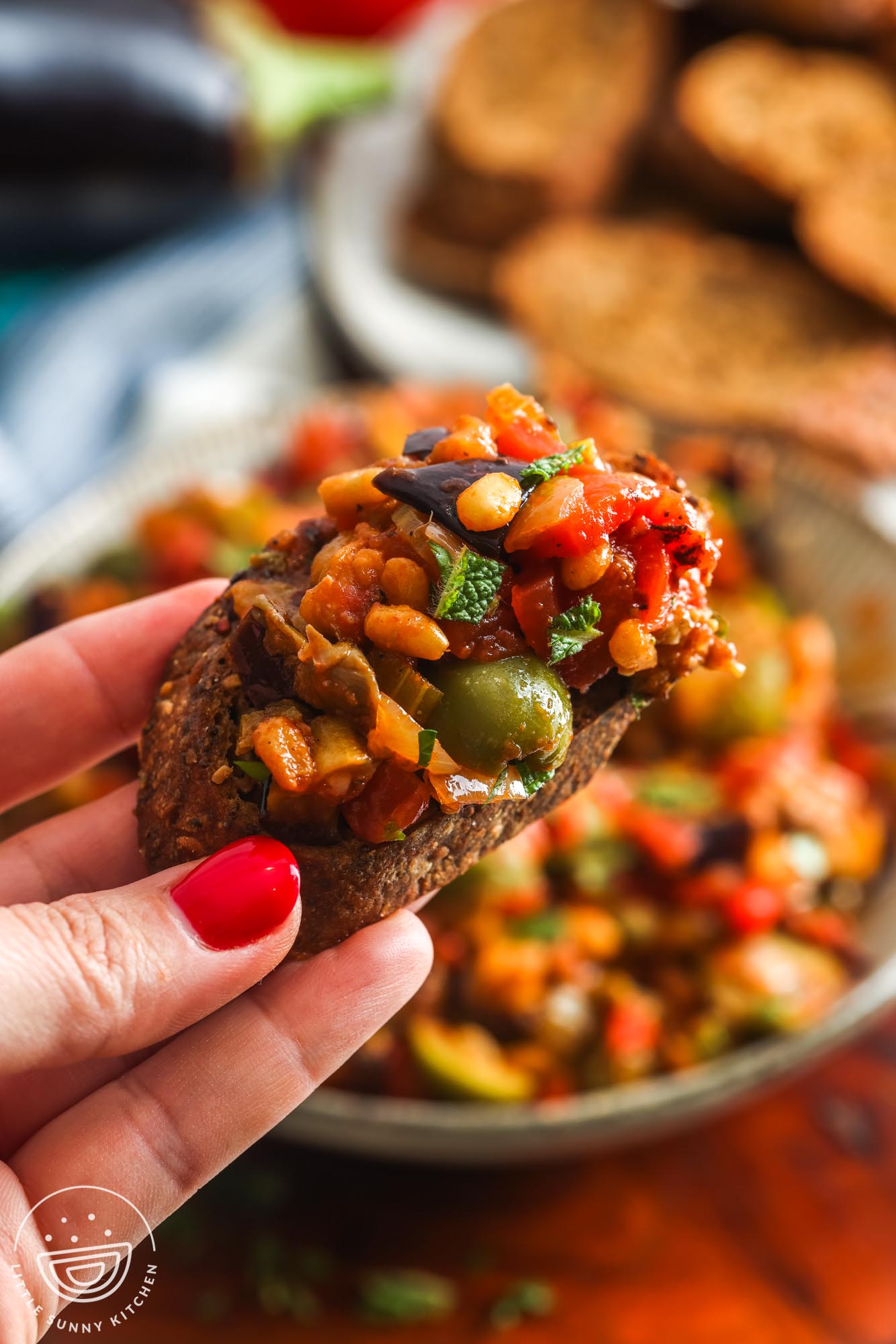 a hand holding a small slice of bread covered in caponata.