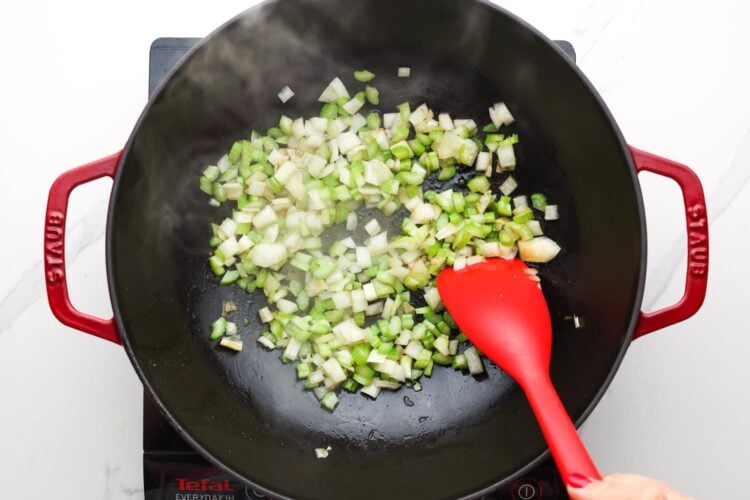 diced onion and celery cooking in a skillet, stirred with a red spatula.