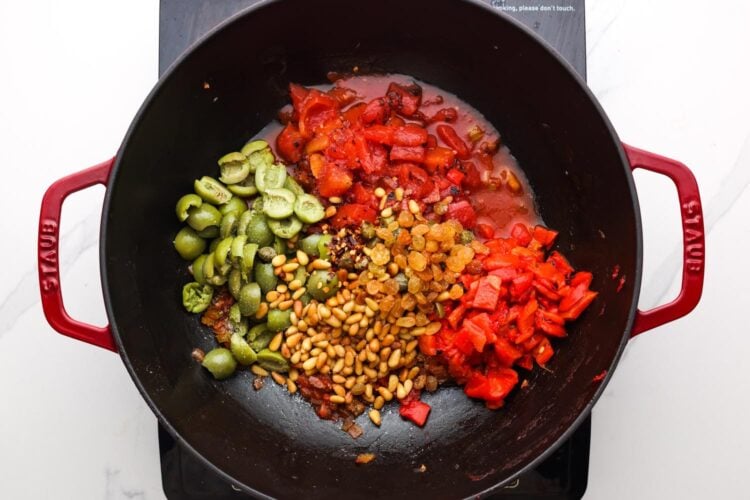 olives, tomatoes, roasted red peppers, raisins and seasonings added to sauteed vegetables and pine nuts to create caponata in a frying pan.