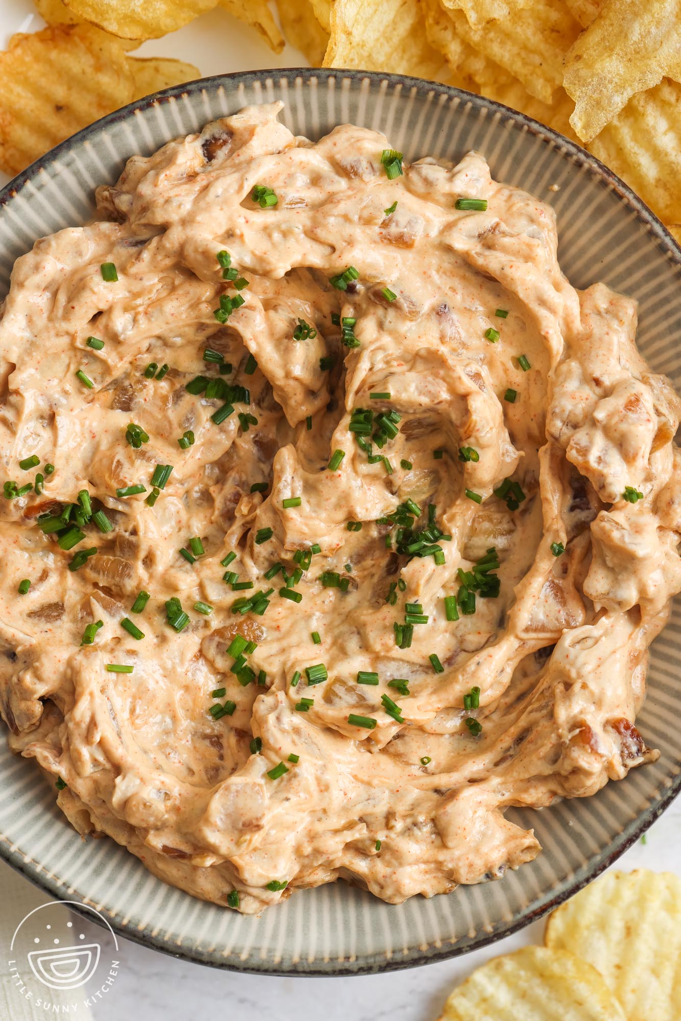 a striped ceramic bowl of creamy caramelized onion chip dip, garnished with chopped chives.