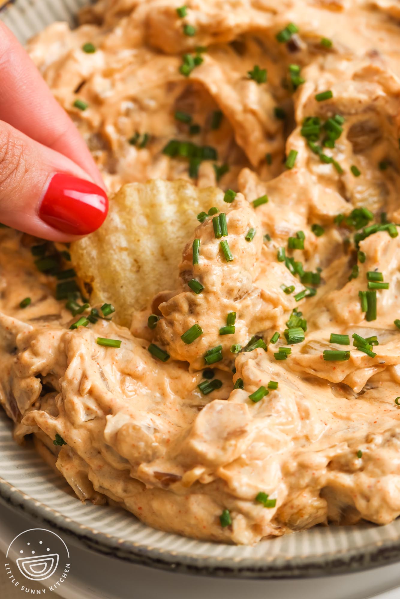 a hand with red fingernail polish dipping a ruffled potato chip into a bowl of creamy onion dip.