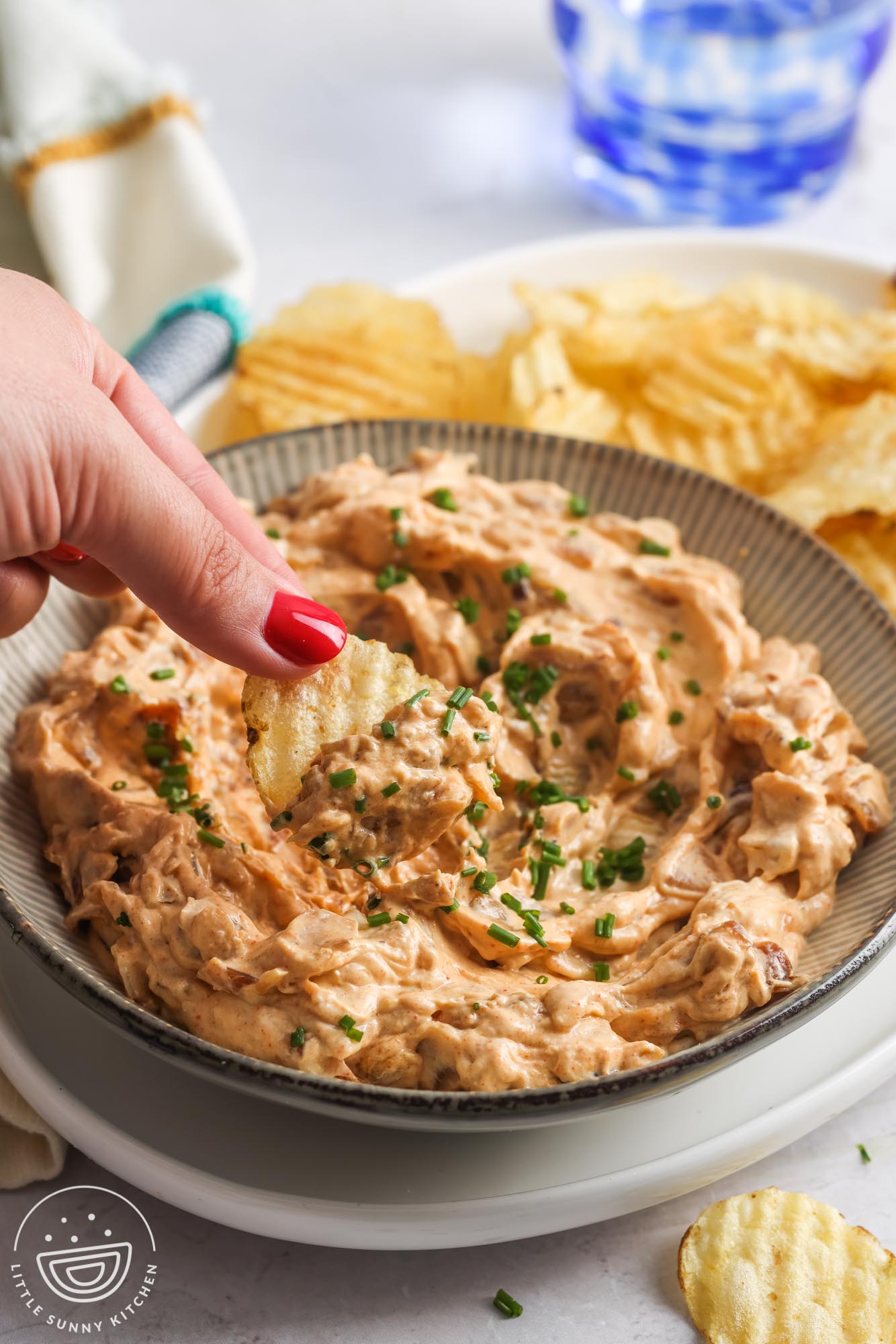 a hand dipping a chip into a bowl of caramelized onion dip. The dip is on a platter with potato chips on the side.