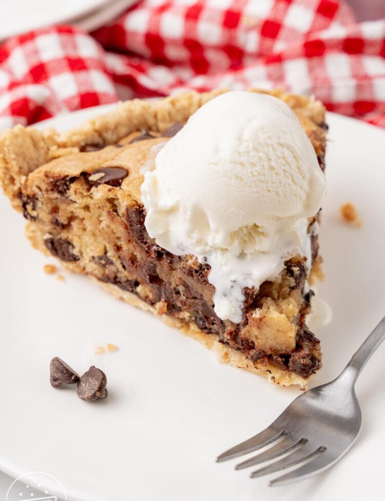 a warm slice of chocolate chip cookie pie topped with a scoop of vanilla ice cream. A fork is on the plate in front of the slice.