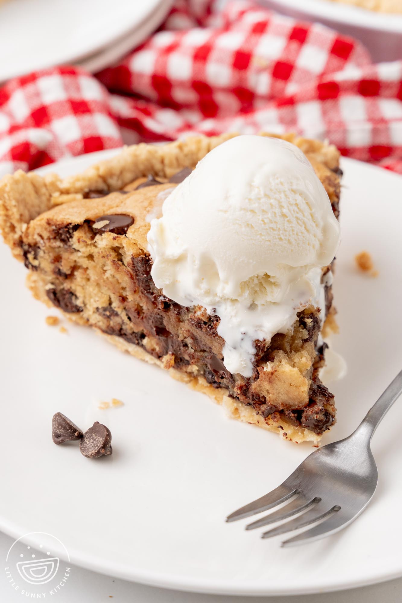 a warm slice of chocolate chip cookie pie topped with a scoop of vanilla ice cream. A fork is on the plate in front of the slice.
