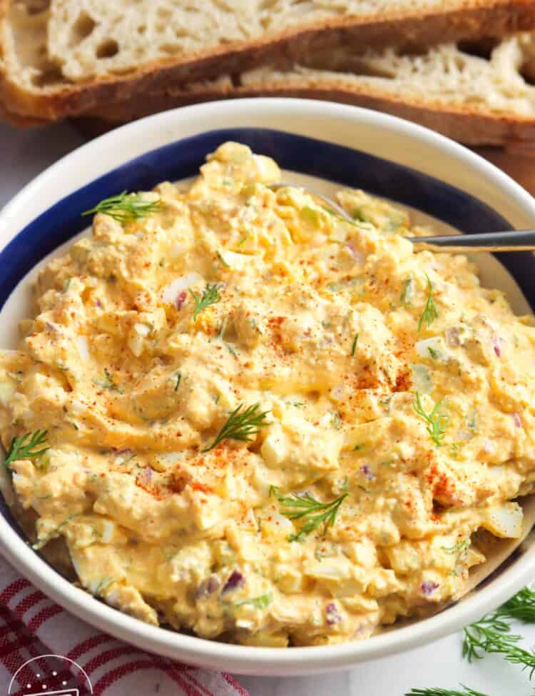 a large bowl of cottage cheese egg salad garnished with dill and paprika, in front of slices of bread.
