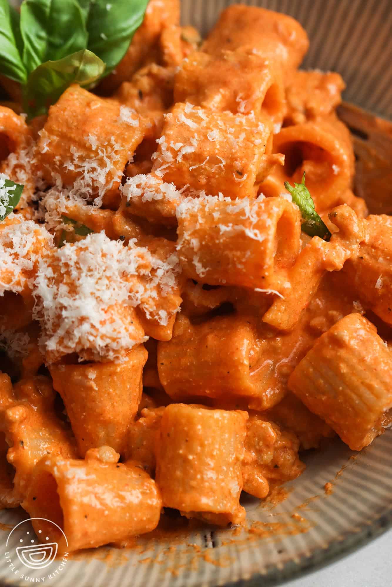 closeup of mezze rigatoni with roasted red pepper cottage cheese sauce on a plate, garnished with basil leaves and grated parmesan.